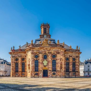 Barocke Ludwigskirche in Saarbrücken mit Sandsteinfassade, Turm und Statuen auf einem Kopfsteinpflasterplatz unter blauem Himmel.