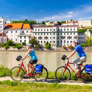 Two cyclists with red bikes and blue panniers riding along the Inn River in Passau, with historic buildings and church towers visible across the water.