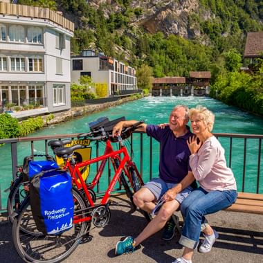 Paar mit Tourenrädern auf Brücke über türkisfarbene Aare in Interlaken, mit traditionellen Gebäuden und bewaldeten Bergen im Hintergrund.