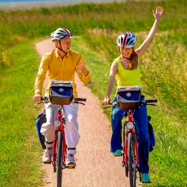 Zwei Radfahrer auf roten Rädern mit Lenkertaschen auf einem Schotterweg durch grüne Felder. Die Frau in Gelb winkt fröhlich.
