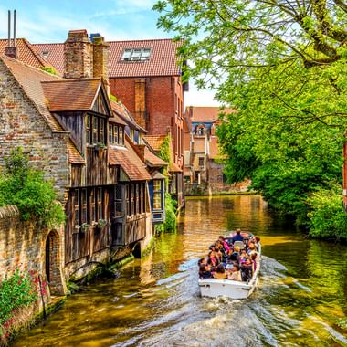 Touristenboot auf einem schmalen Kanal in Brügge, vorbei an historischen Backsteingebäuden mit roten Ziegeldächern und grünen Bäumen.