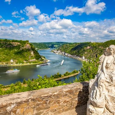 Stone statue at Loreley viewpoint overlooking the Rhine River winding through green hills. Cruise ships sail on the river below under blue sky.