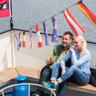 Couple sitting on wooden deck bench of ship Magnifique II with international flag bunting overhead and water in background.