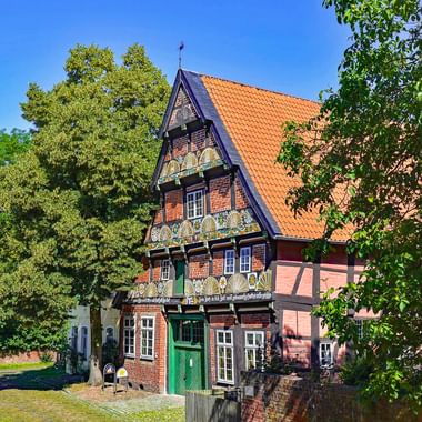 Traditionelles Fachwerkhaus Ackerbürgerhaus in Verden mit bunten Verzierungen, orangefarbenem Ziegeldach und grüner Tür, umgeben von Bäumen.