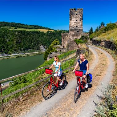 Zwei Frauen fahren mit roten Rädern auf Hangweg nahe mittelalterlicher Turmruine in Kaub. Mosel und Weinberge unter blauem Himmel sichtbar.
