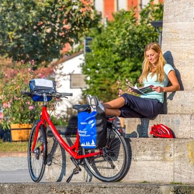 Frau liest Karte auf Steinmauer sitzend neben rotem Tourenrad mit blauen Taschen am historischen Konzilgebäude in Konstanz.