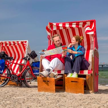 Zwei Radfahrer sitzen in einem rot-weiß gestreiften Strandkorb am Sandstrand in Niendorf und lesen eine Karte. Ein Tourenrad steht daneben.