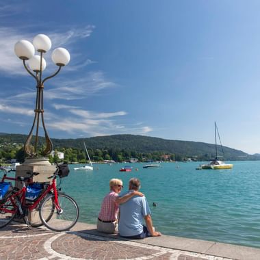 Zwei Radfahrer sitzen an der Uferpromenade in Velden am Wörthersee mit geparkten Fahrrädern. Türkisfarbener See mit Segelbooten und Bergen.