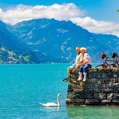 Zwei Radfahrer sitzen auf einer Steinmauer am Brienzersee, ein weißer Schwan schwimmt in der Nähe. Berge und blauer Himmel im Hintergrund.