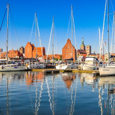 Yachthafen in Stralsund mit Segelbooten im ruhigen Wasser. Historische Backsteingebäude und Kirchtürme säumen die Uferpromenade unter blauem Himmel.