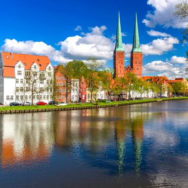 Der Lübecker Dom mit zwei grünen Türmen erhebt sich hinter bunten historischen Gebäuden entlang der Trave unter blauem Himmel mit weißen Wolken.