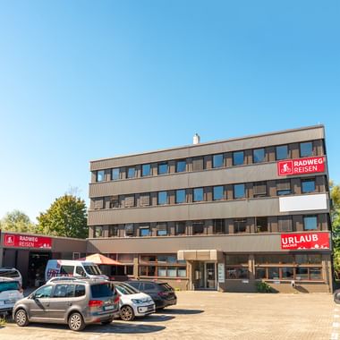 Modernes vierstöckiges Bürogebäude mit Radweg-Reisen Beschilderung in Konstanz. Rote Firmenschilder an der Fassade. Parkplatz mit Autos davor.