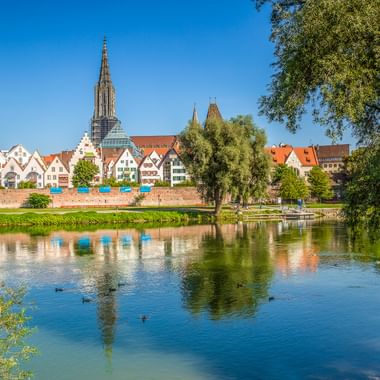 Das Ulmer Münster mit seinem hohen Turm erhebt sich über bunten historischen Gebäuden entlang der Donau. Bäume rahmen die Aussicht und Spiegelungen schimmern im Wasser.