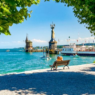 Blick auf den Konstanzer Hafen am Bodensee mit Imperia-Statue, historischem Leuchtturm, Booten auf blauem Wasser und Menschen auf Bank unter grünen Bäumen.