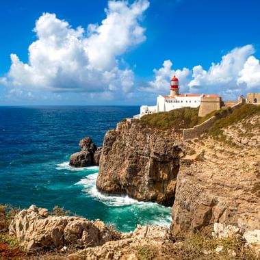 Rot-weißer Leuchtturm an der Festung Cabo de São Vicente auf dramatischen Felsenklippen mit Blick auf den Atlantik unter blauem Himmel mit weißen Wolken.