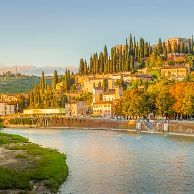 Blick auf Verona mit der Etsch im Vordergrund und einem Hügel mit Zypressen und historischen Gebäuden unter klarem Himmel.