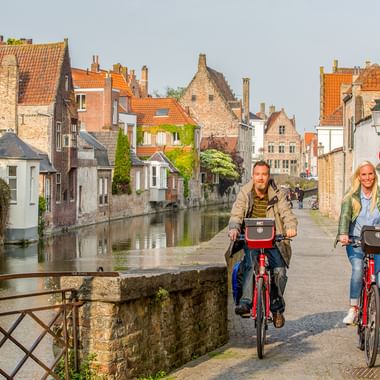 Zwei Radfahrer mit roten Rädern überqueren eine Steinbrücke über einen Kanal in Brügge, mit mittelalterlichen Backsteingebäuden und orangefarbenen Ziegeldächern im Hintergrund.