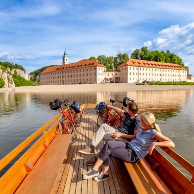 Zwei Radfahrer mit Rädern auf Holzboot überqueren die Donau. Kloster Weltenburg mit roten Ziegeldächern am gegenüberliegenden Ufer sichtbar.