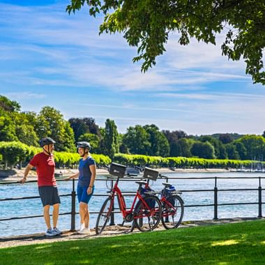 Zwei Radfahrer mit Helmen stehen mit ihren Rädern an der Seepromenade in Konstanz, mit grünen Bäumen und Booten im Hintergrund.