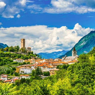 Gemona del Friuli eingebettet in grüne Hügel mit mittelalterlichem Turm auf einem Hügel, Kirchturm und dramatischen Berggipfeln unter blauem Himmel.