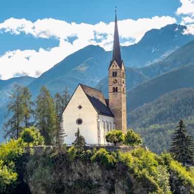 Weiße Kirche mit hohem spitzem Turm auf einem Hügel in Scuol, umgeben von grünen Bäumen mit dramatischen Alpenbergen im Hintergrund.