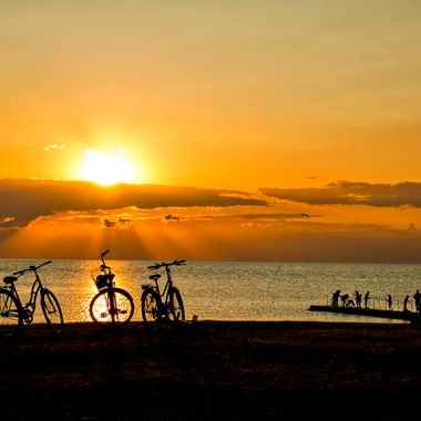 Drei Fahrräder als Silhouetten vor goldenem Sonnenuntergang über dem Meer in Torekov. Menschen versammeln sich auf einem Steg im Hintergrund.