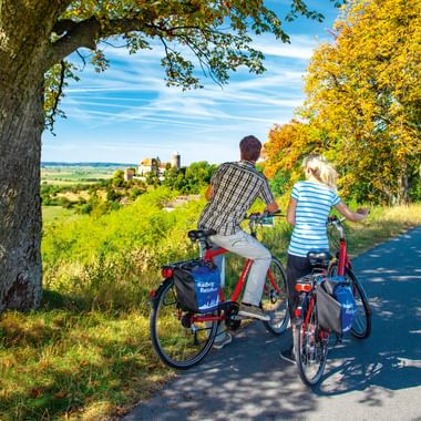 Zwei Radfahrer mit Packtaschen machen Pause auf einem Weg mit Blick auf das Altmühltal mit Burgruine, Herbstbäumen und Landschaft.