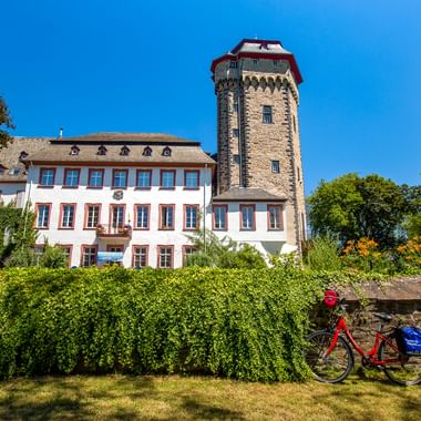Schloss Martinsburg in Lahnstein mit Steinturm und weißem Gebäude, umgeben von grünen Hecken. Ein rotes Fahrrad steht im Vordergrund auf einer Wiese.