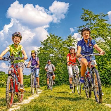 Fünf Radfahrer mit Helmen fahren auf einem Grasweg. Zwei Kinder führen, gefolgt von drei Erwachsenen. Grüne Bäume und blauer Himmel mit weißen Wolken im Hintergrund.
