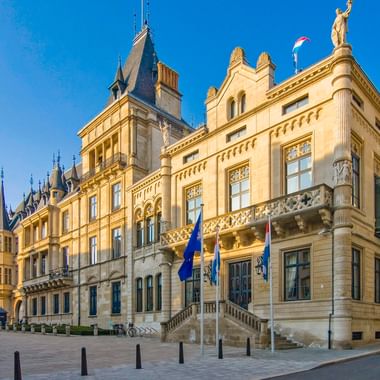 Großherzoglicher Palast in Luxemburg mit verzierter beiger Fassade, dekorativen Balkonen und Flaggen. Das historische Gebäude steht auf einem Platz.