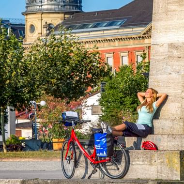Frau in Radkleidung entspannt an Steinsäule mit rotem Tourenrad daneben. Historisches Konzilgebäude im Hintergrund in Konstanz sichtbar.
