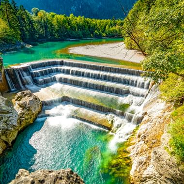 Stufenwasserfall am Lechfall in Füssen mit türkisfarbenem Wasser, das über fünf Stufen in eine felsige Schlucht fällt, umgeben von grünem Wald.