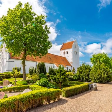 Weiße Stubberup Kirche mit rotem Ziegeldach und Turm, umgeben von gepflegten Hecken, Blumenbeeten und grünen Bäumen unter blauem Himmel auf Fünen.