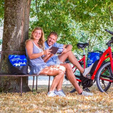 Zwei Radfahrer sitzen auf einer Bank unter einem Baum in Stein am Rhein und schauen auf ein Tablet. Zwei rote E-Bikes mit blauen Taschen stehen daneben.