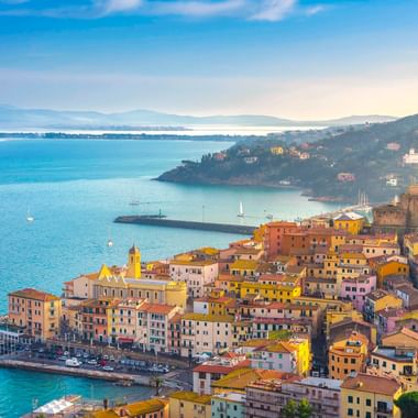 Luftaufnahme von Porto Santo Stefano mit bunten Gebäuden am Hang, Hafen mit Booten und blauem Mittelmeer in der Toskana.