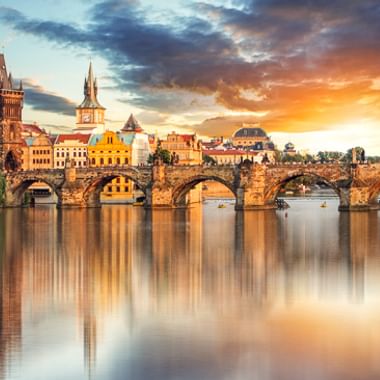 Karlsbrücke über die Moldau in Prag bei Sonnenuntergang, mit historischen Türmen und bunten Gebäuden, die sich im ruhigen Wasser spiegeln.