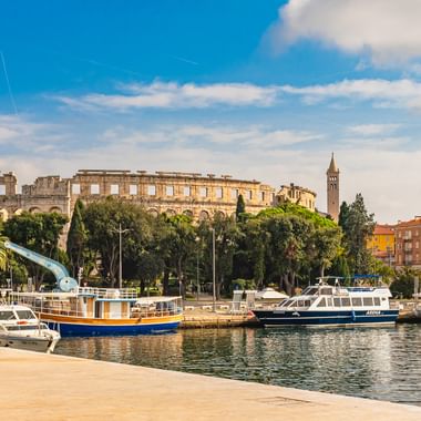 Hafen von Pula mit vertäuten Booten auf ruhigem Wasser. Römisches Amphitheater und historische Gebäude auf dem Hügel mit Bäumen unter blauem Himmel.