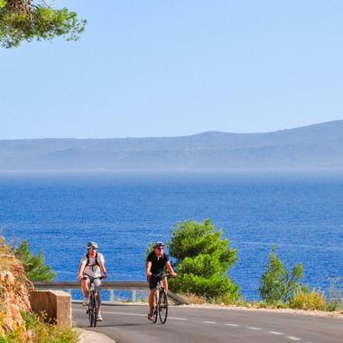 Zwei Radfahrer auf einer Küstenstraße auf der Insel Brač, Kroatien, mit blauer Adria und fernen Bergen unter klarem Himmel.