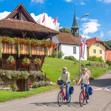 Zwei Radfahrer auf einer Dorfstraße in Böhningen, Schweiz, vorbei an einem blumengeschmückten Holzbau und einer weißen Kirche mit grünem Turm.