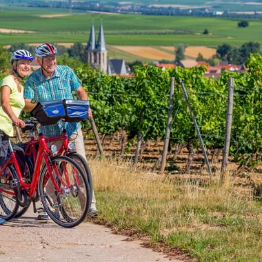 Zwei Radfahrer mit Helmen und rotem Fahrrad pausieren auf Weg neben Weinberg in Flonheim, Hessen. Kirchtürme und Felder im Hintergrund.