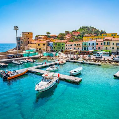 Hafen von Giglio Porto mit Booten im türkisfarbenen Wasser, bunten Häusern am Ufer und einem historischen Turm links.
