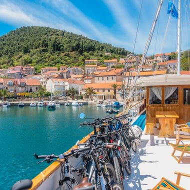 Reihe von Fahrrädern auf Holzschiffsdeck mit Liegestühlen, Blick auf türkisfarbenen Hafen und Küstenstadt Korčula mit roten Dächern.