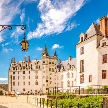White stone Château des Ducs de Bretagne in Nantes with towers and turrets under blue sky. Courtyard with visitors and historic lantern visible.