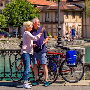 Älteres Paar mit Fahrrädern auf einer Brücke in Interlaken, das auf Sehenswürdigkeiten zeigt. Türkisfarbener Kanal und historische Gebäude im Hintergrund.