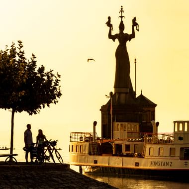 Silhouette der Imperia-Statue im Hafen Konstanz bei Sonnenaufgang mit Raddampfer und zwei Personen mit Fahrrädern auf der Kopfsteinpflaster-Promenade.