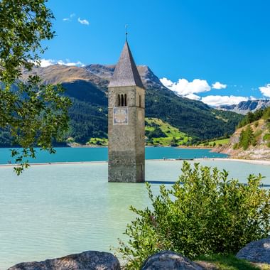 Historischer Kirchturm ragt aus dem türkisfarbenen Wasser des Reschensees bei Graun, Südtirol, mit Bergen und grüner Vegetation.