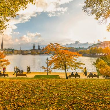 Herbstansicht der Hamburger Binnenalster mit goldenem Laub, Menschen auf Bänken und der Stadtsilhouette mit Kirchtürmen bei Sonnenuntergang.
