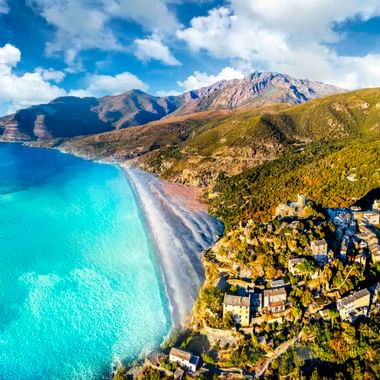 Luftaufnahme des Dorfes Nonza auf grünem Hang über türkisfarbenem Mittelmeer und dunklem Strand, mit Bergen im Hintergrund unter blauem Himmel.