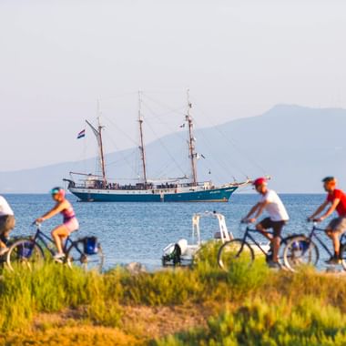 Vier Radfahrer fahren entlang eines Küstenwegs in der Toskana mit einem Dreimaster auf dem Meer und Bergen im Hintergrund.