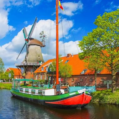 Historische Windmühle mit weißen Flügeln neben einem Kanal in Großefehn, Ostfriesland. Ein buntes Boot liegt vor Backsteingebäuden mit orangen Dächern.